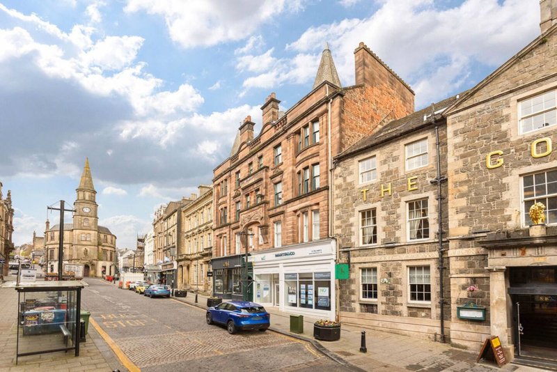 Street view of historic stone buildings with shops, parked car, and church tower in background