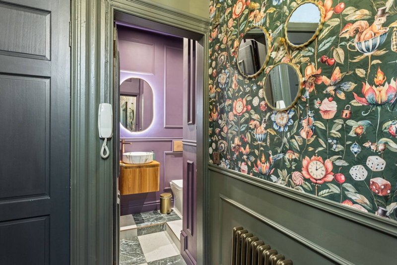 Hallway with open bathroom door, floral wallpaper, mirrors, and sage green painted walls