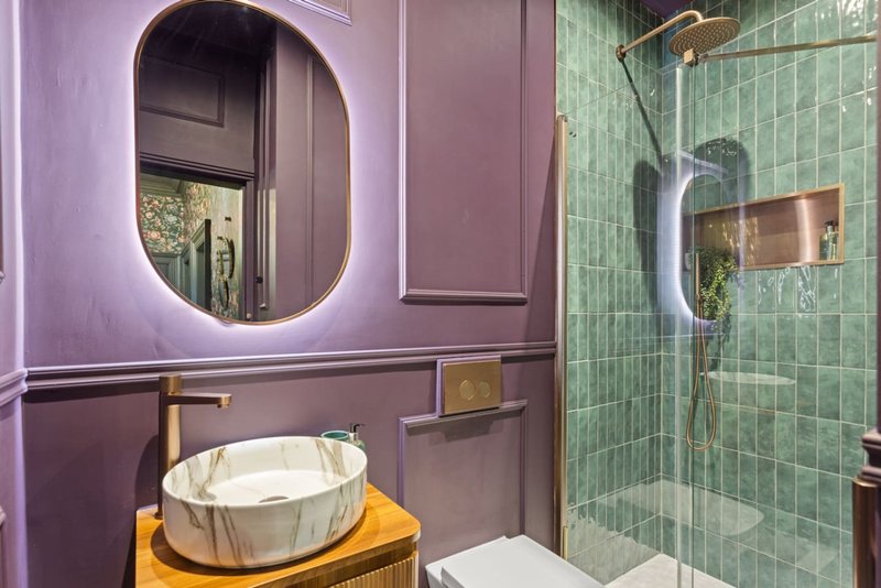 Modern bathroom with purple walls, round lit mirror, vessel sink and green tile shower