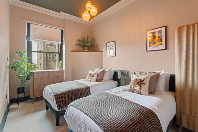 Twin bedroom with two beds, cushions, window with blinds, framed wall art, and potted plant