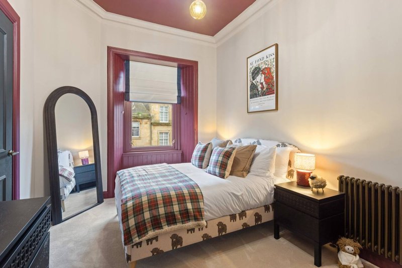 Bedroom with double bed, plaid bedding, wooden nightstands, mirror and window with burgundy frame