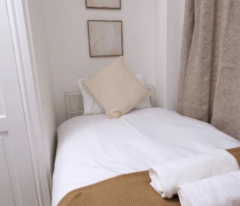 Compact bedroom with single bed, cream pillow, white bedding and beige curtains.
