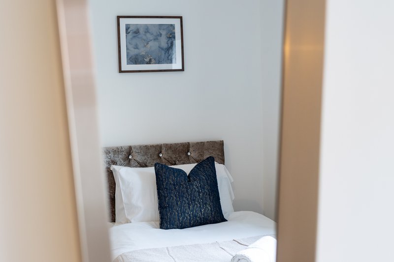 Compact bedroom with single bed, gray headboard, white bedding, dark blue cushion, framed wall art