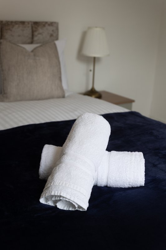 Bedroom with rolled white towels on dark blue bedding, lamp and headboard visible