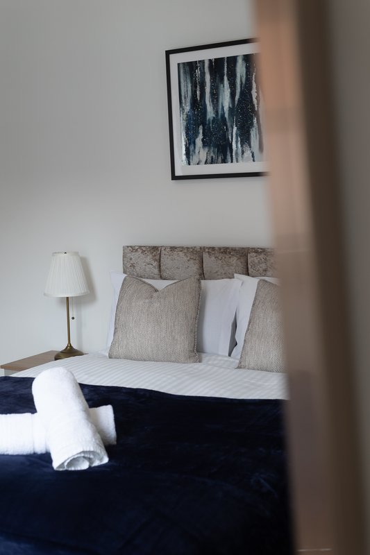 Bedroom with double bed, grey headboard, cushions, bedside lamp and framed wall art