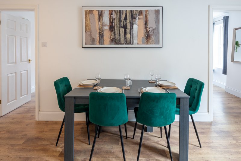 Dining room with dark table, four green upholstered chairs, framed artwork and white doors