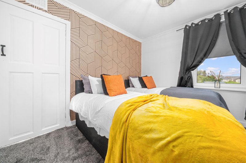 Modern bedroom with double bed, yellow blanket, orange pillows, geometric headboard, and window with curtains