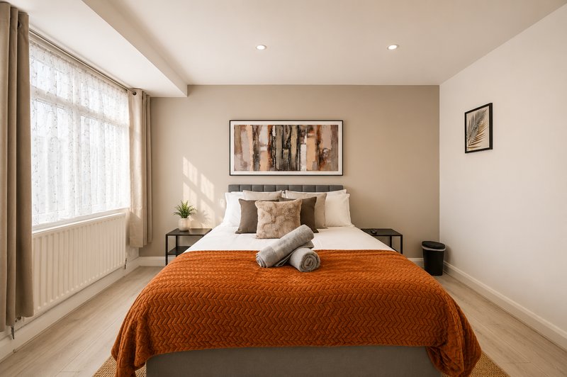 Bright bedroom with orange bedspread, neutral walls, framed artwork, and large window with curtains