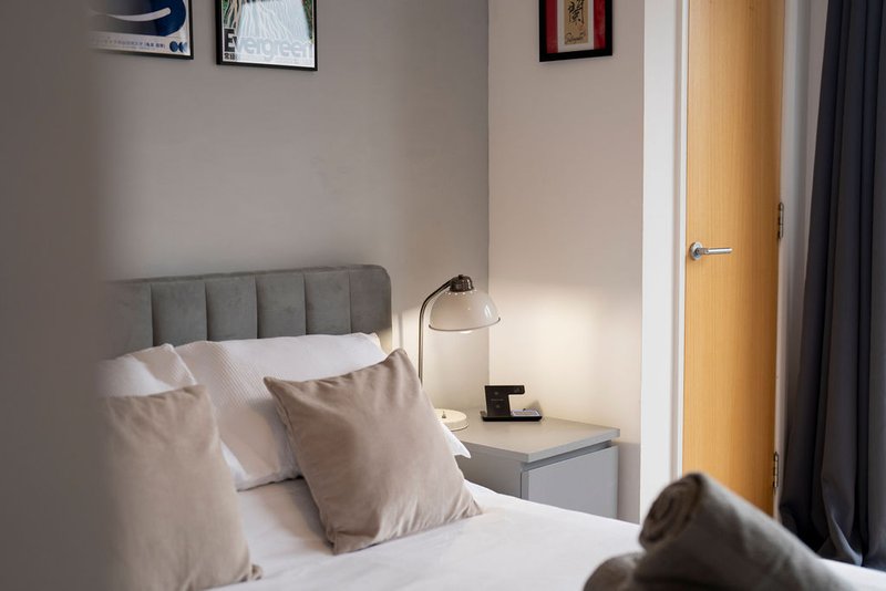 Modern bedroom with grey upholstered headboard, white bedding, beige cushions and bedside lamp