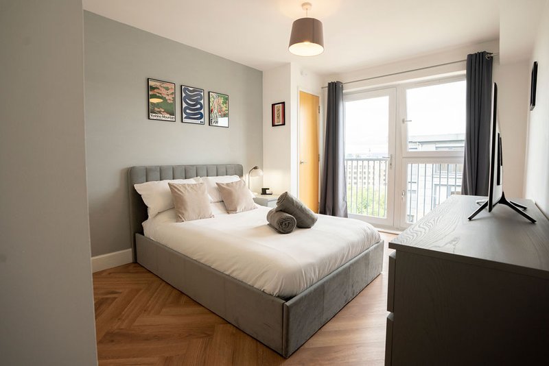 Bright modern bedroom with gray upholstered bed, wooden floor, pendant light, and balcony access