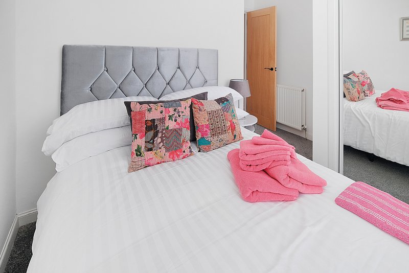 Bedroom with grey upholstered headboard, white bedding, colourful cushions and pink throws