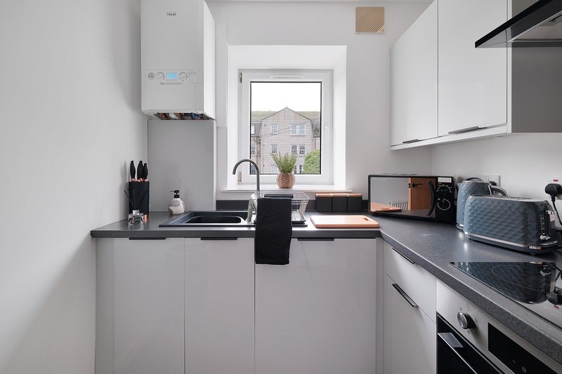 Modern white kitchen with dark countertops, gas hob, window view, and stainless steel appliances