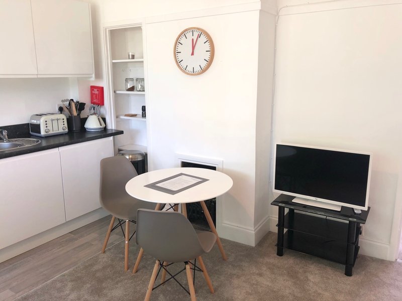 Open-plan kitchen and living area with dining table, chairs, wall clock and TV
