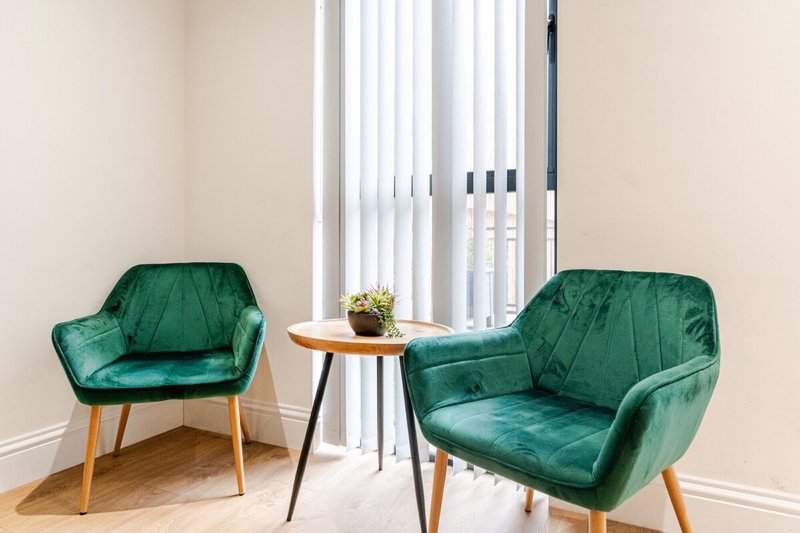 Two green velvet armchairs with wooden legs flanking small side table near vertical blinds