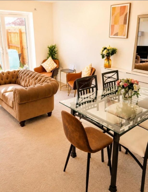 Living room with tan leather sofa, glass dining table, upholstered chairs and floral arrangements