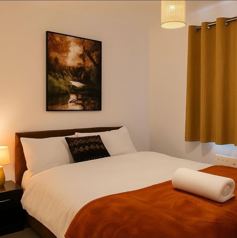 Bedroom with double bed, white bedding, rust-colored throw, framed landscape artwork, and gold curtains