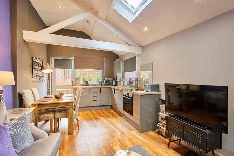 Open-plan kitchen and living area with wooden flooring, grey cabinetry, dining table, and TV