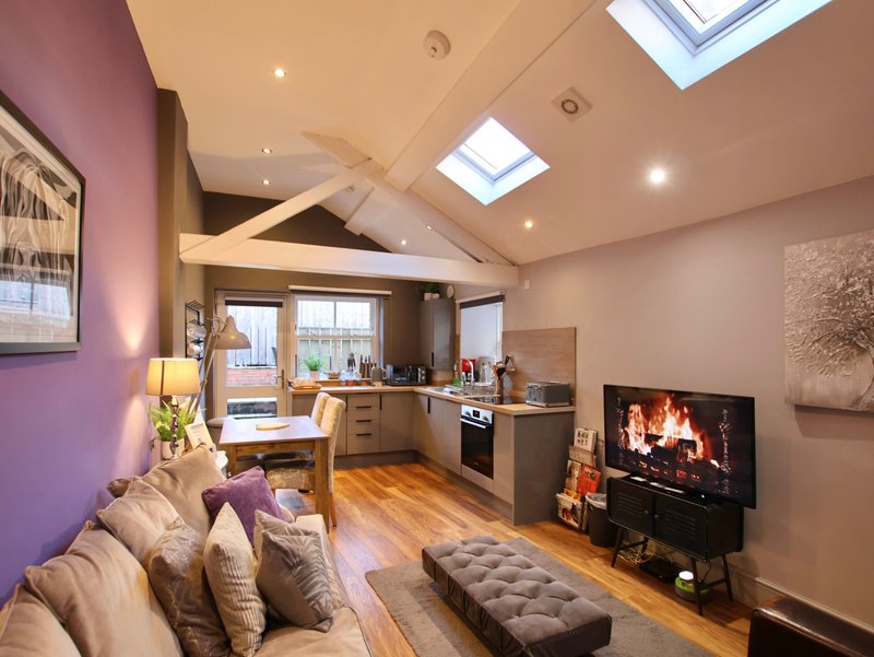 Open-plan living room and kitchen with purple accent wall, wooden beams, skylight and fireplace