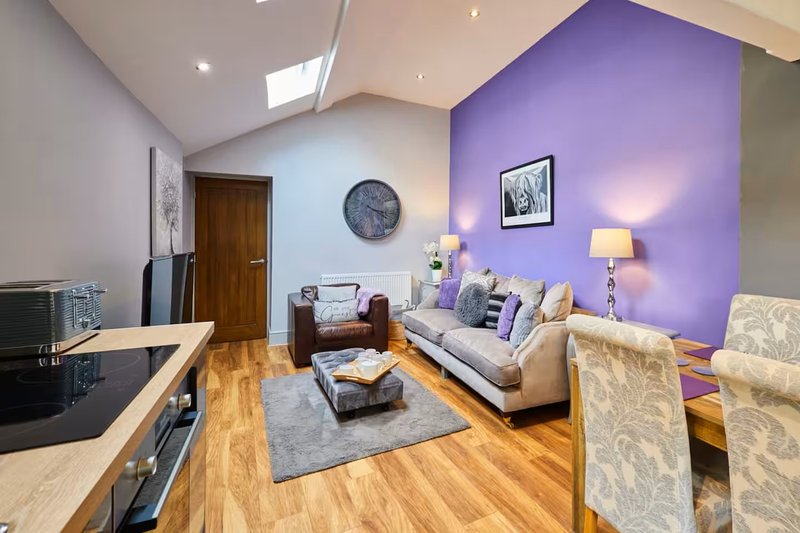 Open-plan living room with purple accent wall, cream sofa, wooden flooring and kitchen area