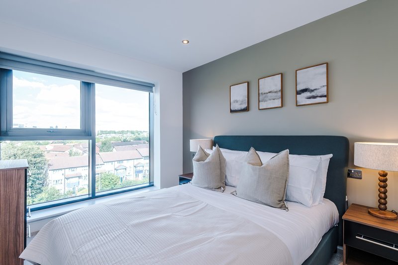 Modern bedroom with double bed, grey headboard, white bedding, large windows overlooking city