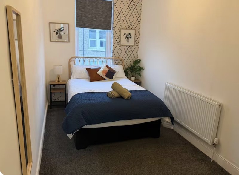Bedroom with double bed, navy bedding, wooden headboard, bedside table and window with blinds