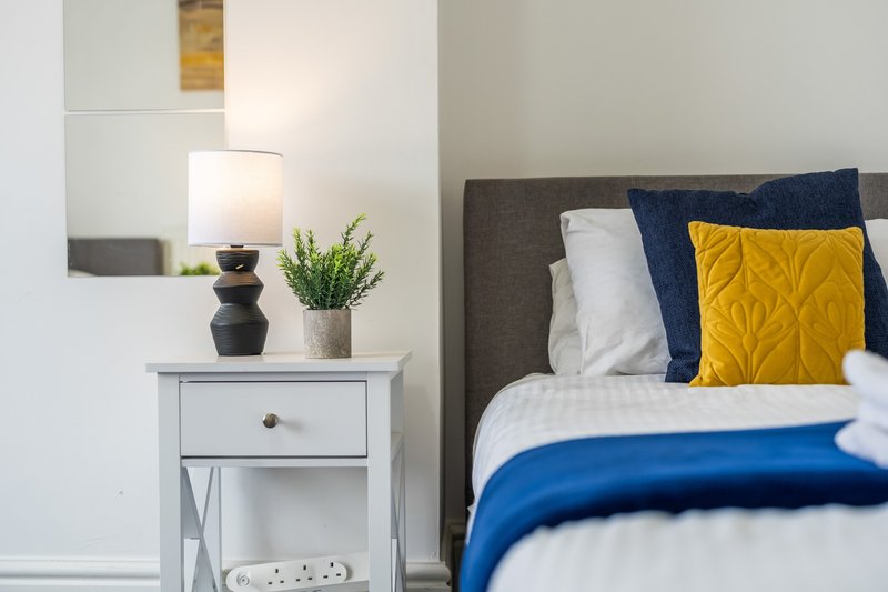 Modern bedroom with grey upholstered bed, yellow and navy cushions, white bedside table with lamp