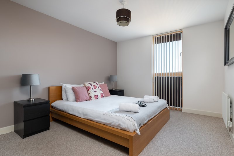 Bright bedroom with wooden bed, pink pillows, bedside table, lamp and vertical blinds.