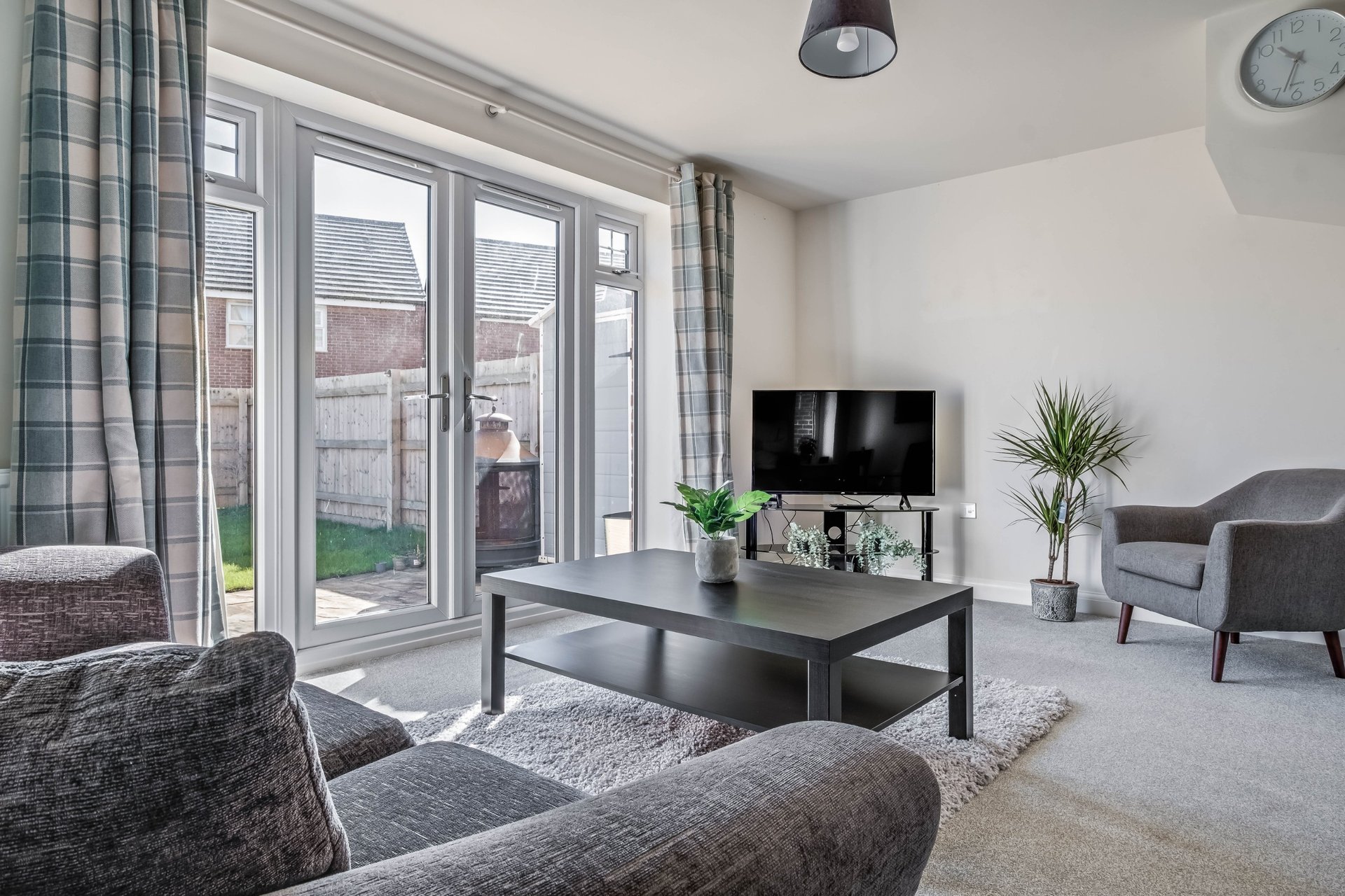Bright living room with grey upholstered furniture, black coffee table, TV, and patio doors overlooking garden