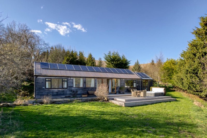 Modern single-story house with solar panels, large windows, wooden deck, and spacious garden surrounded by trees