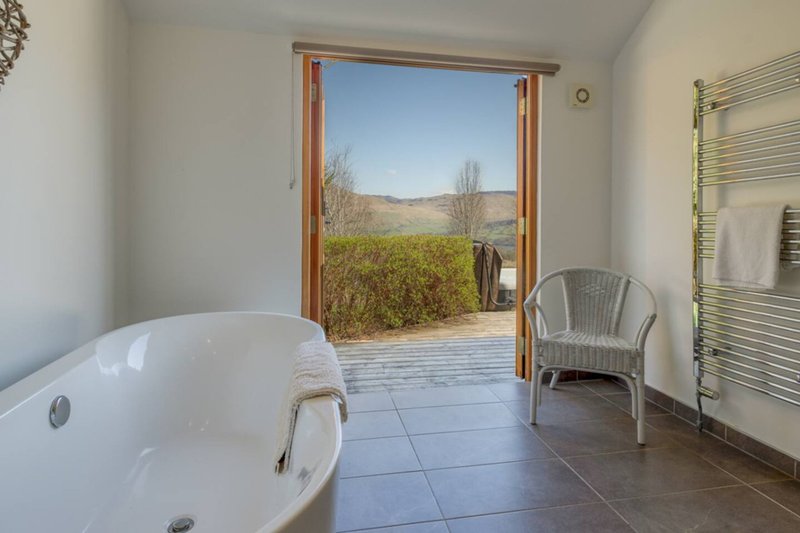 Bathroom with freestanding white bathtub, wicker chair, and open doors revealing countryside landscape view