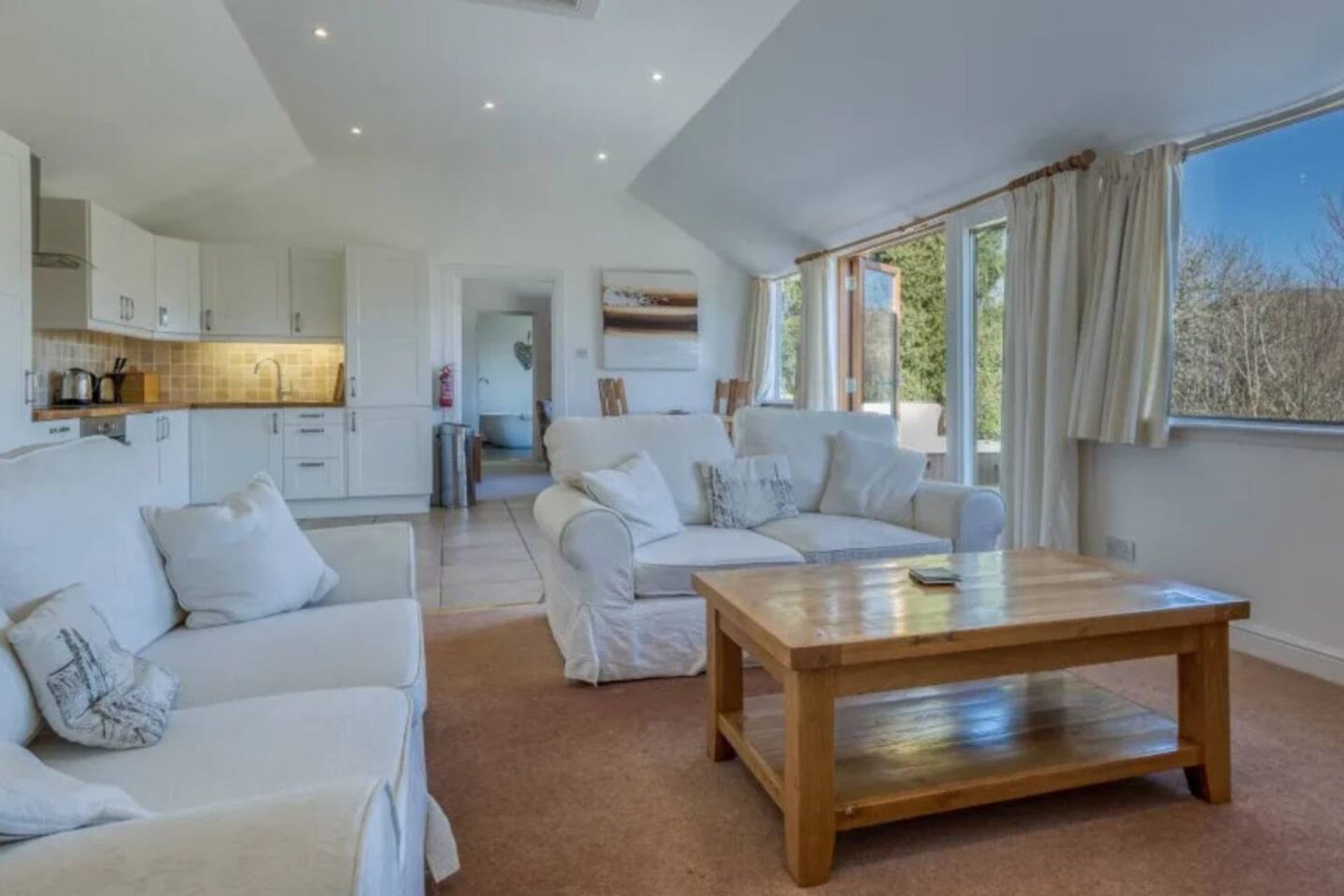 Open-plan living room with cream upholstered sofas, wooden coffee table, kitchen area, and large windows with garden views