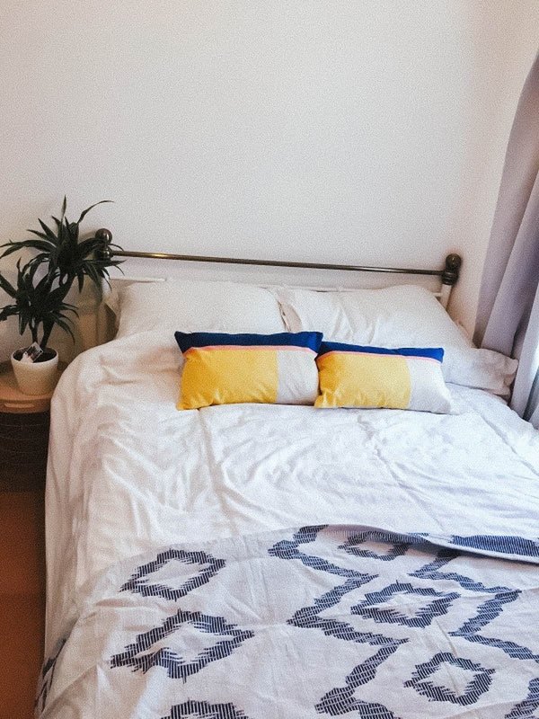 Bedroom with double bed, white bedding, yellow and white pillows, patterned throw blanket, potted plant and curtains