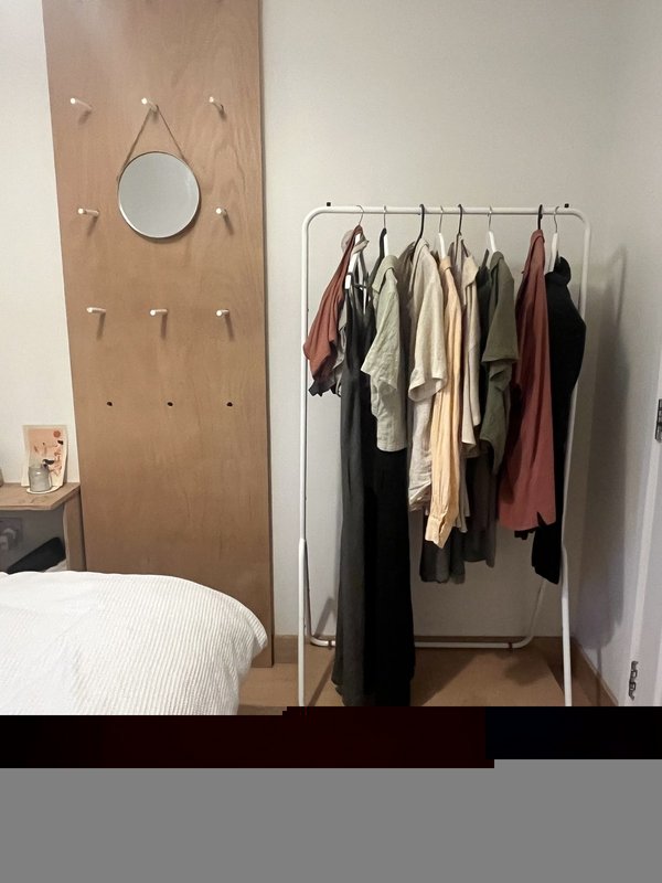 Bedroom corner with pegboard wall, round mirror, clothing rack, and white bed