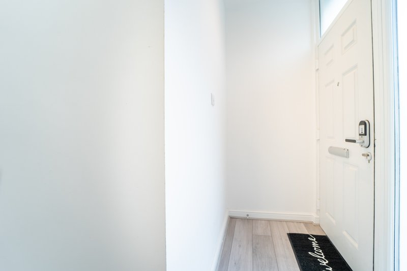 Narrow entryway with white walls, wooden flooring, and a black welcome mat