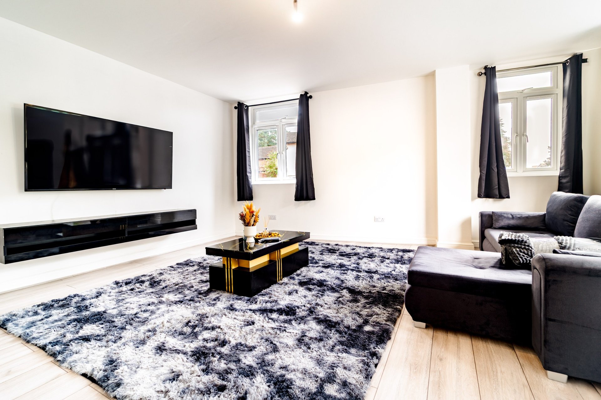 Modern living room with wall-mounted TV, black fireplace, patterned rug, dark sofa and windows