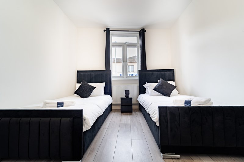 Twin bedroom with black upholstered beds, white bedding, dark cushions, and window with curtains.
