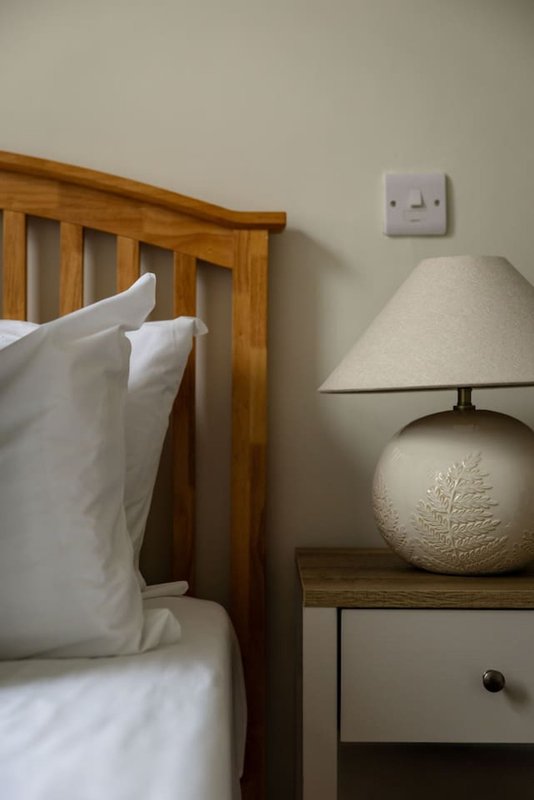 Bedroom corner with wooden bed frame, white pillows, bedside table, table lamp and wall outlet