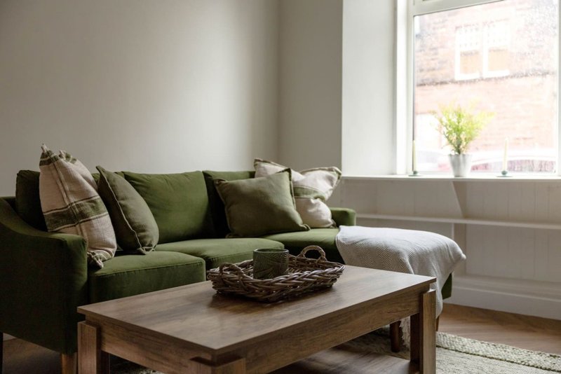 Living room with green upholstered sofa, wooden coffee table, decorative cushions, and window with city view