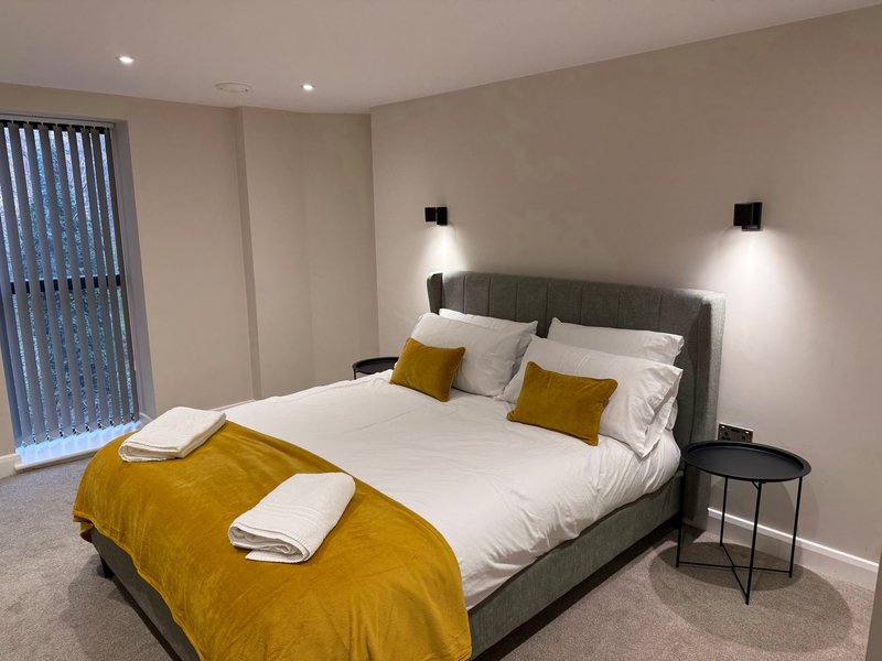 Modern bedroom with double bed, yellow throw, white bedding, grey headboard and wall lights