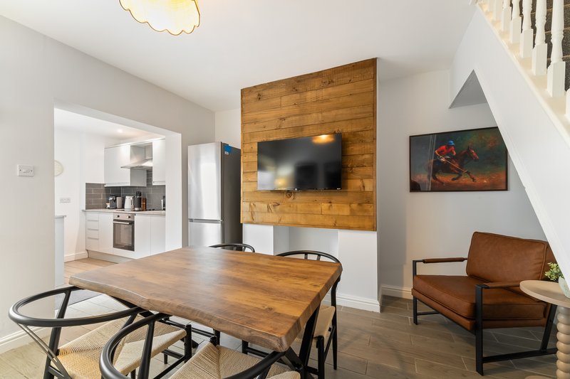 Open-plan living and dining area with wooden table, leather armchair, TV on wood wall, and kitchen visible through doorway