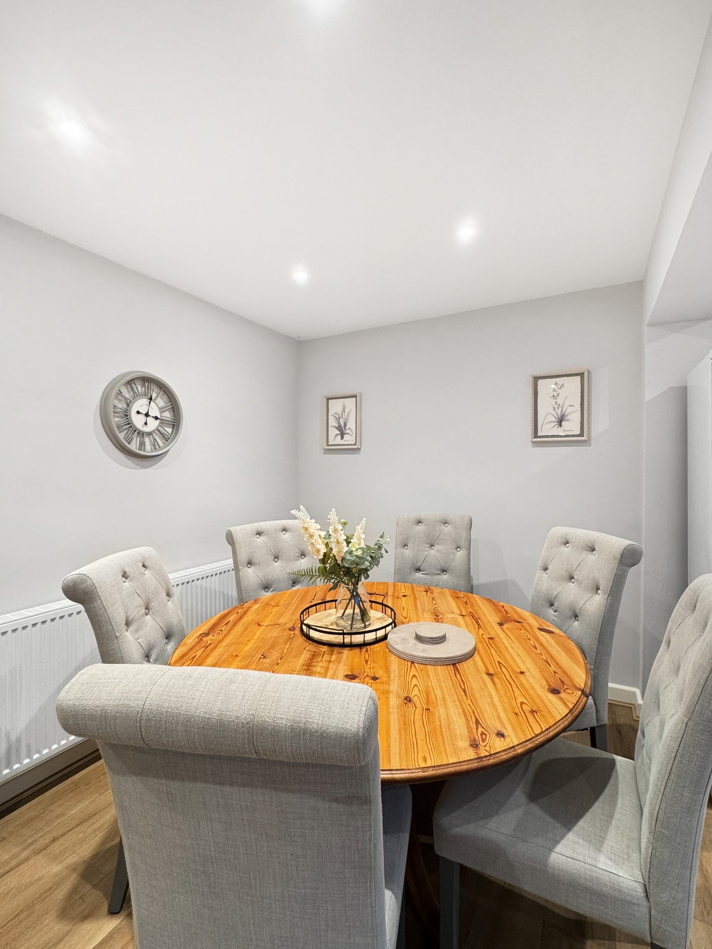 Dining room with round wooden table, upholstered chairs, wall clock and framed botanical prints