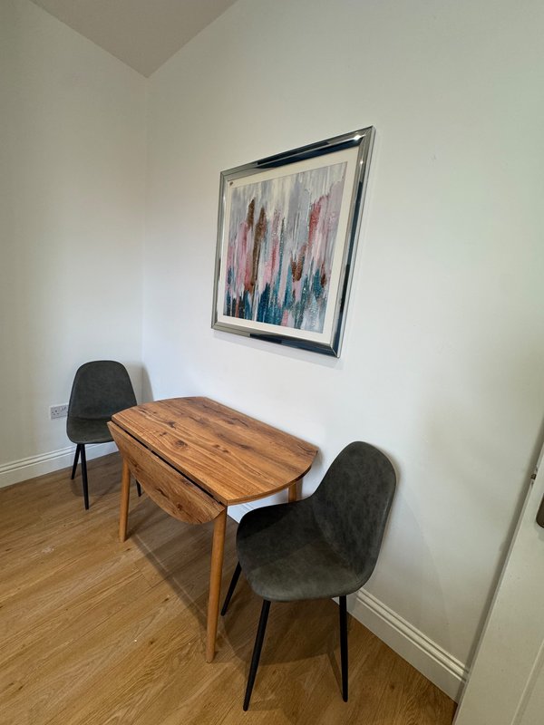 Dining area with wooden table, two dark chairs, and framed abstract artwork on white wall
