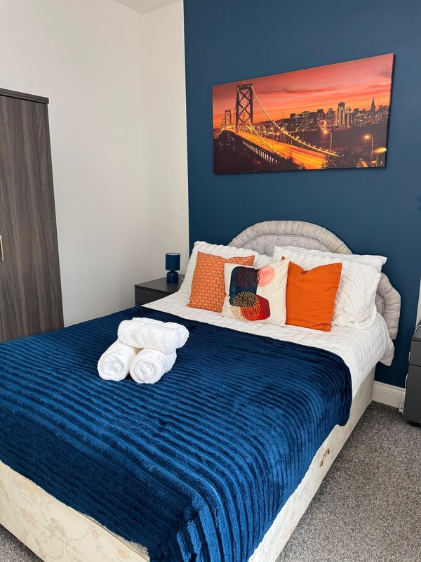 Bedroom with double bed, blue bedding, white pillows, orange cushions and wall art of bridge at sunset