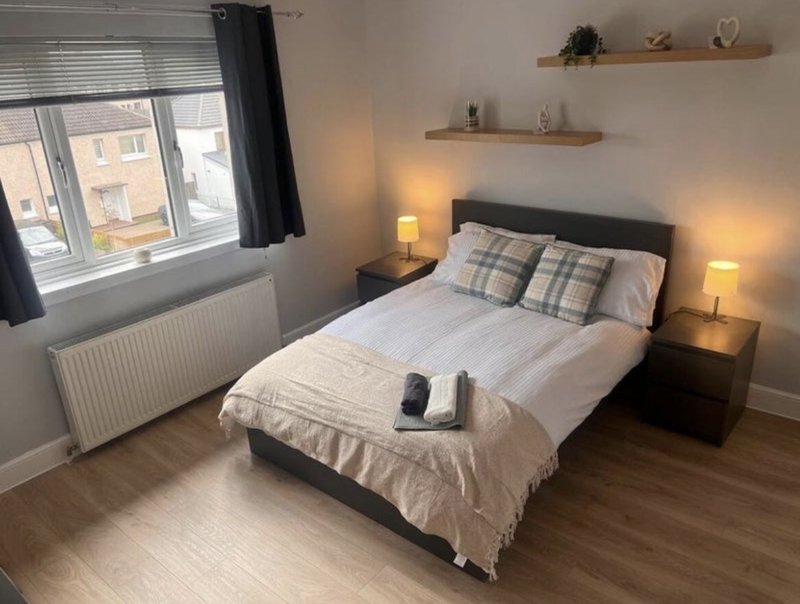 Modern bedroom with double bed, plaid pillows, bedside lamps, and window with city view