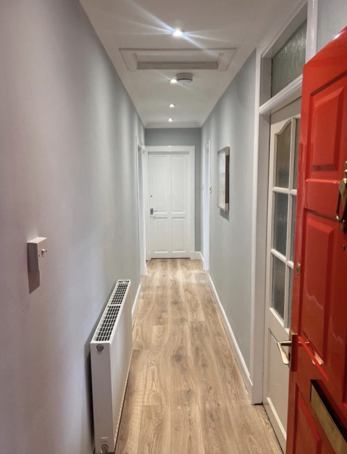 Long narrow hallway with white walls, wooden flooring, red door on right side