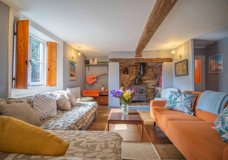 Open-plan living room with exposed brick fireplace, orange and neutral furnishings, wooden beams and window seating