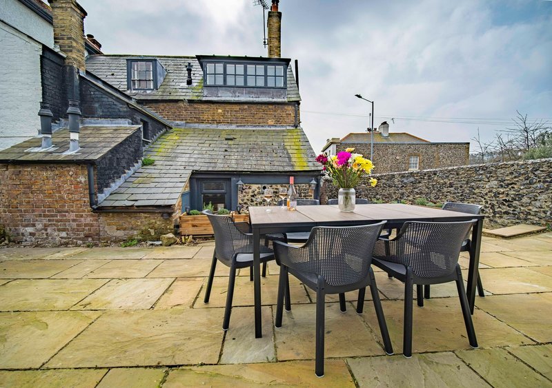 Outdoor patio with dining table, chairs, and colorful flowers overlooking brick house and garden