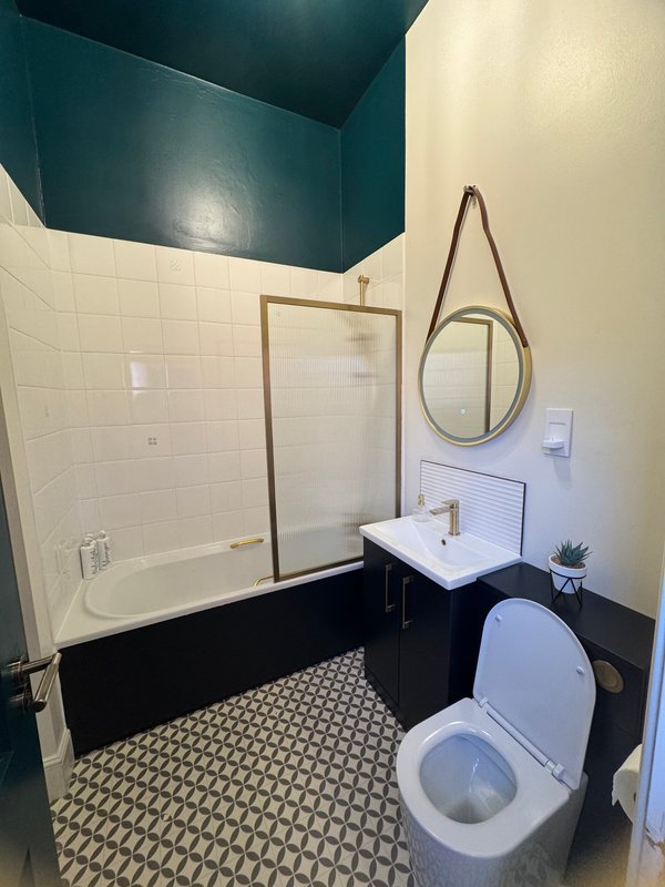 Modern bathroom with teal ceiling, white subway tile walls, black paneling, patterned floor tiles, toilet, sink and round mirror
