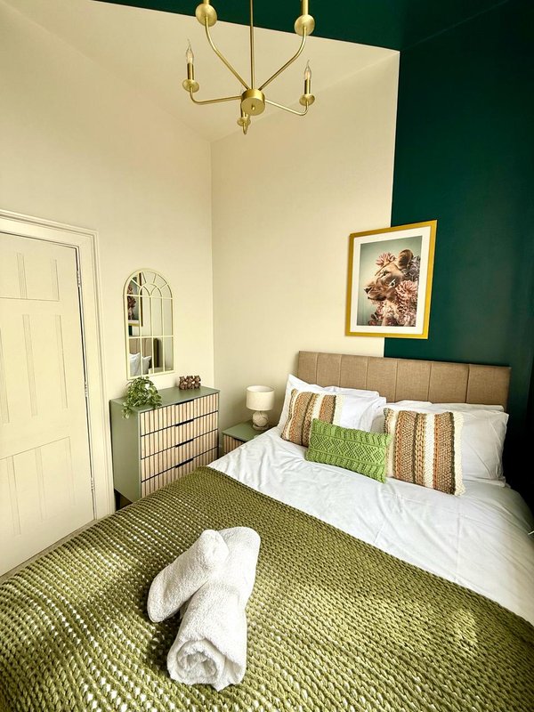 Bedroom with double bed, green accent wall, brass chandelier, and rolled towel on carpet floor