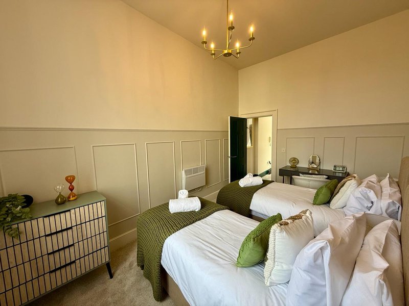 Neutral-toned bedroom with two beds, green bedding, wicker furniture and brass chandelier